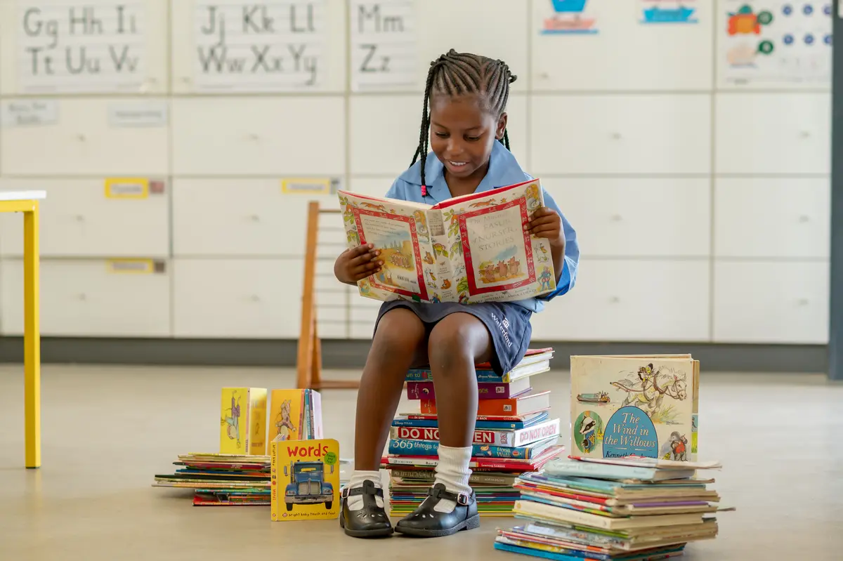A girl sitting on a stack of books, reading, in a bright classroom