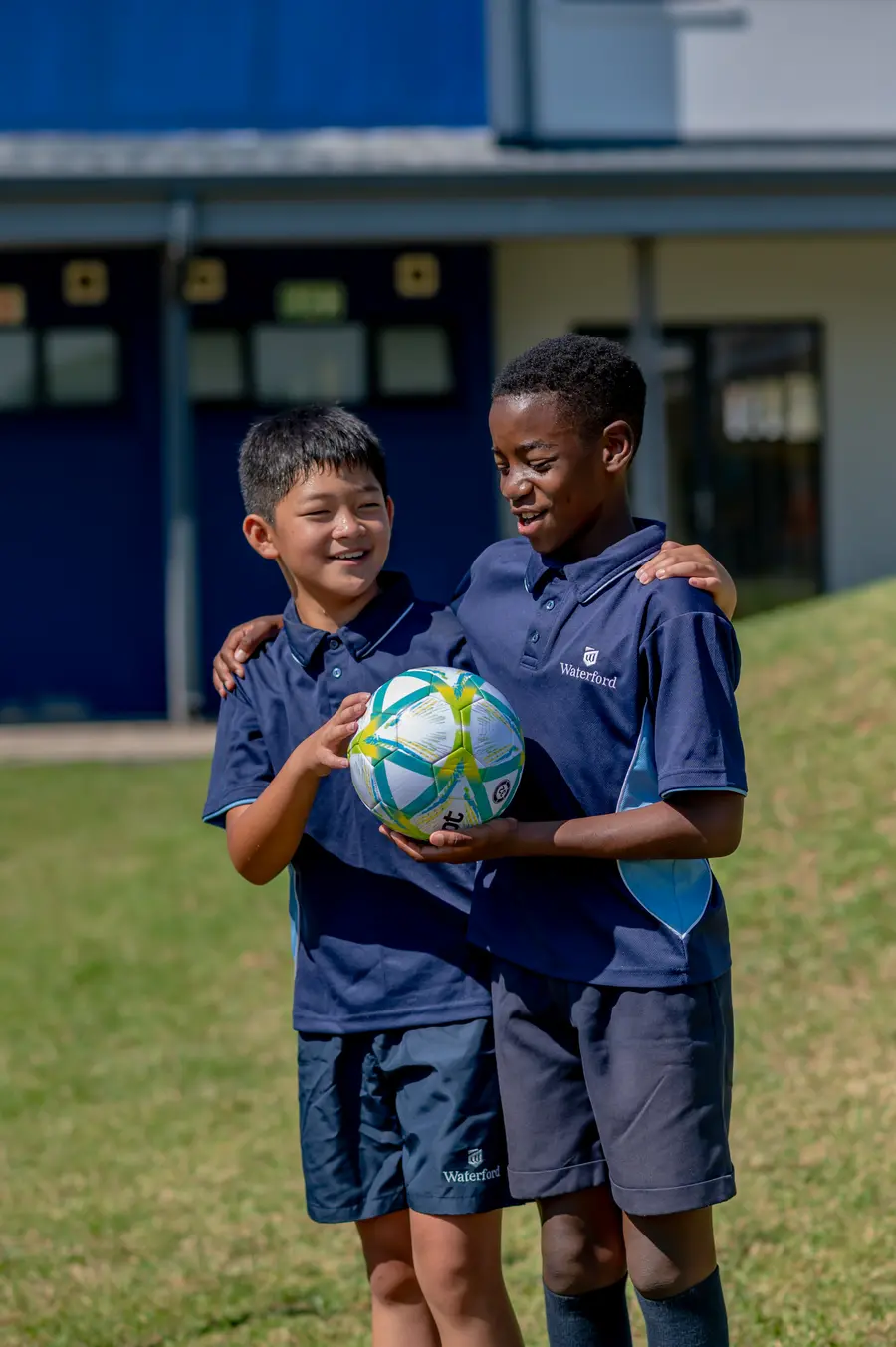Two boys in sports kit standing together on the grass, one holding a soccer ball